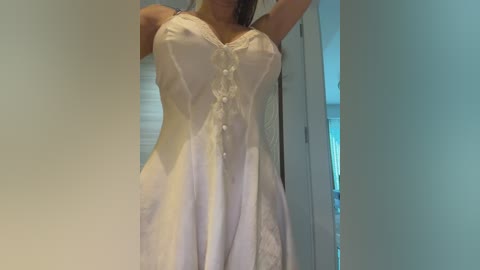 A video of a woman in a white, lacy nightgown with buttons down the front, standing in a dimly lit room with light-colored walls. Her arms are raised, and her breasts are partially visible.