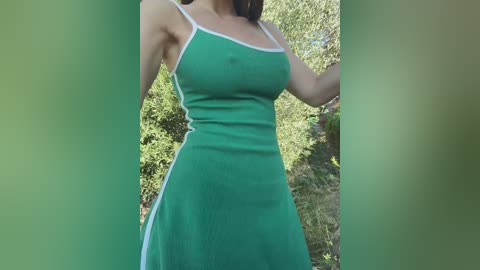Video of a woman in a green, sleeveless dress with white trim, standing outdoors in a sunlit garden with green foliage in the background.