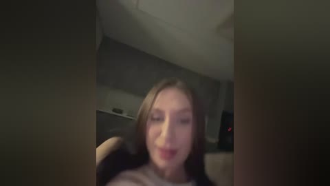 A blurred video of a young woman with light skin and straight, shoulder-length brown hair, wearing a white top, seated in a car with dim lighting.