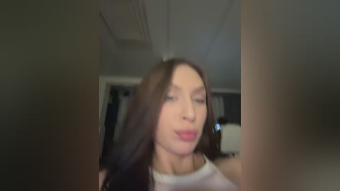 Video of a fair-skinned woman with long, dark hair, wearing a white tank top, smiling seductively in a dimly lit room with visible ceiling panels and blurred background.