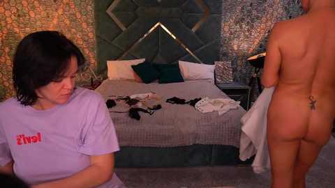 Video of a nude woman with a tattoo on her back, standing in a bedroom with a green, patterned wallpaper, a bed with white sheets, and a purple-clad woman in the foreground.