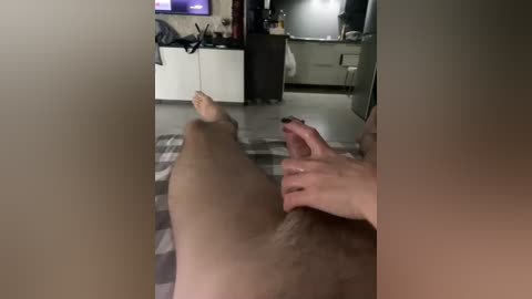 Video of a person lying on a bed with legs spread, their hands holding a circumcised, erect penis. Background shows a modern kitchen with cabinets and a TV.