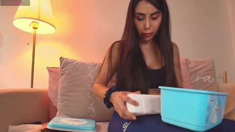 Video of a young woman with long black hair, wearing a black tank top, sitting on a beige couch. She is focused on pouring yogurt into a blue bowl. A beige lamp and pink pillows are in the background.