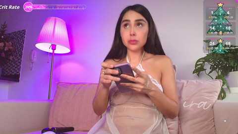 Video of a young woman with long black hair, wearing a sheer white dress, playing with a smartphone in a cozy living room with a lamp, Christmas tree, and a pink sofa.