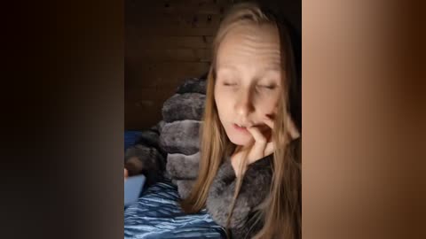 Video of a young girl with long blonde hair, lying on a bed with a blue blanket, eyes closed, hand on her mouth, in a dimly lit room with wooden walls.