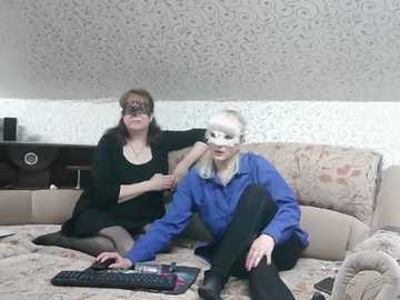 Video of two women on a beige couch, one wearing a black mask, the other a blue mask; floral wallpaper background.