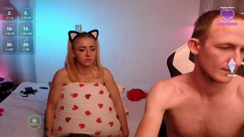 Video of a blonde woman in a cat hat, wearing a red heart pillow, and a shirtless man with short hair, sitting in a gaming chair in a dimly lit room.