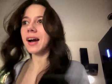 Video of a young woman with shoulder-length dark hair, wearing a gray top, smiling and speaking, indoors with a TV and dim lighting.