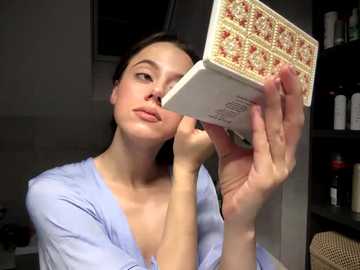 A video shows a young Caucasian woman with fair skin and brown hair, wearing a light blue button-down shirt, examining a box of expensive chocolates with intricate designs. The background is a dimly lit room with shelves filled with bottles and a woven hat.