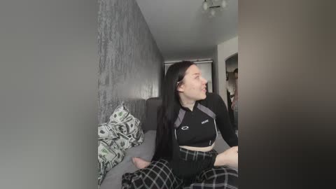 A video of a young woman with long dark hair, wearing a black crop top and plaid pajama bottoms, sitting on a gray couch in a dimly lit room with a textured gray wall and a patterned pillow.