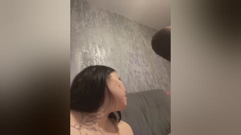 Video of a young Asian woman with long black hair, fair skin, and a small tattoo on her neck, sitting indoors against a textured, metallic-gray wall.
