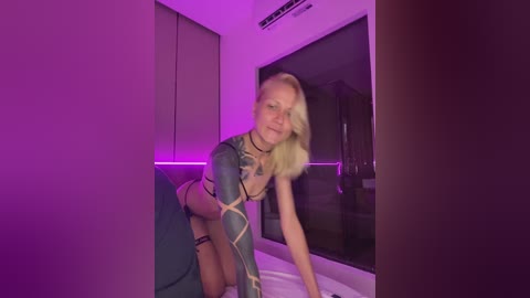 Video of a blonde, fair-skinned woman with medium breasts, wearing a blue and gold tattooed bodysuit, posed on a bed in a dimly lit room with purple lighting, modern decor.