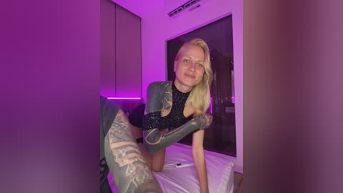 Video of a blonde woman in a black glittery top and camouflage-patterned thigh-high boots, posing on a bed with purple lighting in a modern bedroom.