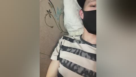 Video of a person with light skin, wearing a black mask, a striped shirt, and a white pillow. They are lying on a beige wall with floral patterns.