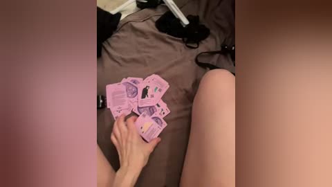 A video shows a light-skinned person's hand holding pink banknotes on a bed. The background includes a brown sheet, black lingerie, and a white object, possibly a phone charger.