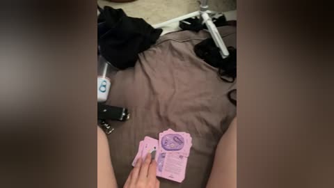Video of a person's legs on a bed with a brown sheet, holding pink pills and keys. Clothes and a bicycle are partially visible in the background.