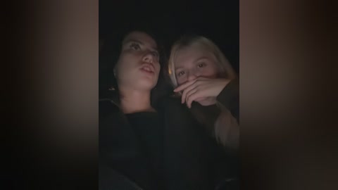 Video of two women, one with dark hair, the other with blonde hair, both with fair skin, sitting close together, partially obscured by shadows, creating a sense of intimacy.