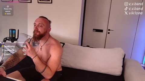 Video of a shirtless, muscular, bearded man with tattoos, sitting on a white couch, holding a phone, in a modern living room with white walls and framed art.