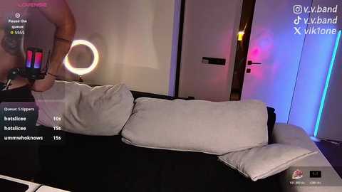 A video of a man wearing a black leather mask, holding a white pillow, in a modern bedroom with a glowing ring light, white walls, and a door.
