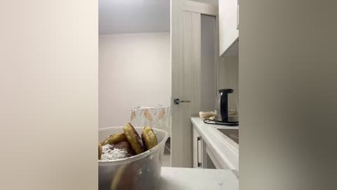 A video of a modern, minimalist kitchen with a white countertop, stainless steel appliances, and a clear bowl containing fried potatoes and a white cloth.