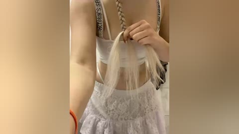 A video of a woman with fair skin, braided platinum blonde hair, and a white lace dress. She's holding the ends of her hair, and the background is blurred, suggesting an indoor setting.