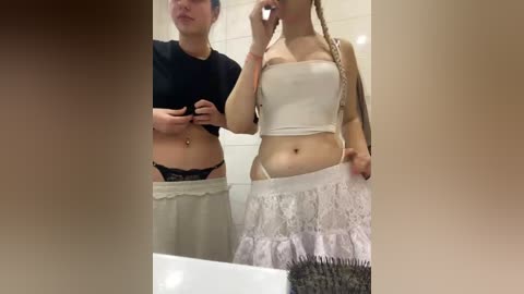 Video of two young women in a bathroom; one wearing a black crop top and white skirt, the other in a white tube top and lace skirt.