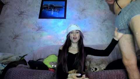 Video of a young woman with long dark hair wearing a white hat and black top, seated on a purple couch in a dimly lit room.