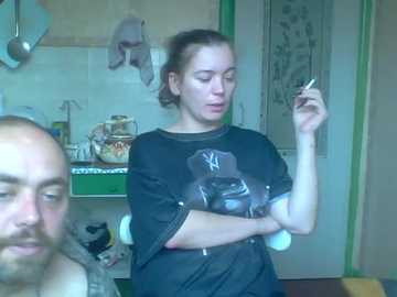 A video of a young woman with light skin and dark hair in a ponytail, wearing a black t-shirt with a Yankees logo, smoking a cigarette indoors. The background includes a cluttered kitchen with a whiteboard, utensils, and a door.