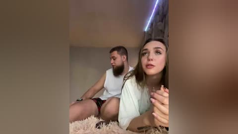 Video: A bearded man with short hair, wearing a white tank top and shorts, sits on a beige carpet. A woman with long brown hair, in a light shirt, sits beside him, holding a glass.