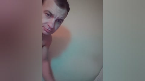 A video shows a young man with short brown hair, wearing no shirt, peering through a narrow gap between two walls. The background is blurred and dimly lit, creating an intimate, slightly voyeuristic atmosphere.