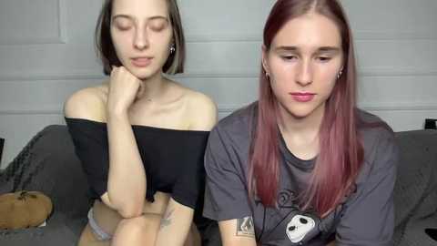 Video of two young women with fair skin, one with short dark hair and off-shoulder black top, the other with long red hair and gray t-shirt, sitting on a gray couch, looking sad.