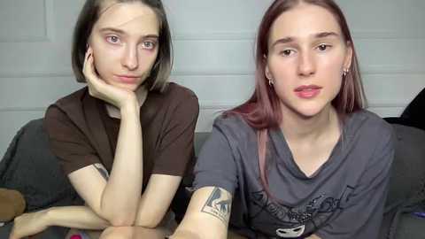 Video of two young women with pale skin, one with shoulder-length brown hair, the other with long, straight reddish-brown hair. They sit side-by-side on a couch, both wearing casual grey T-shirts, looking introspective.