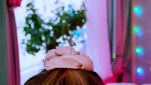 Video of a woman with light brown hair, wearing a pink headband, lying face down on a massage table. Background features a blurred potted plant and colorful string lights.