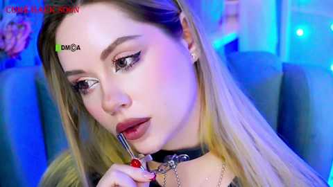 Video of a light-skinned, blonde woman with sharp makeup, including dark eyeliner and bold red lipstick, applying lip gloss. She wears a black choker and has a green \"DMGA\" watermark.