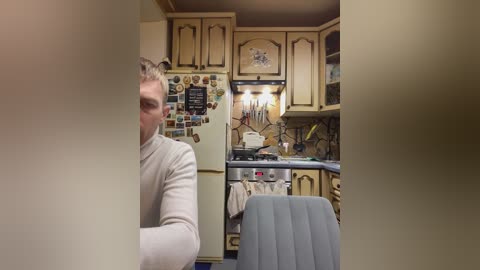 A video of a kitchen featuring a man in a beige sweater, a beige refrigerator covered in photos, light wood cabinets, and a grey chair.