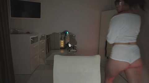 Video of a dimly lit kitchen with a woman in white lingerie, revealing her buttocks, standing near a counter. Appliances and a microwave are visible in the background.