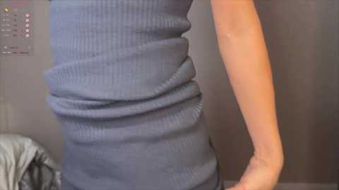 A video of a woman's torso in a tight, ribbed, blue dress, showcasing her slim figure, standing indoors with a gray wall and a glimpse of a white bed sheet in the background.