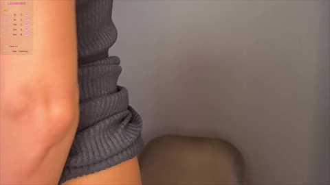 A video of a person's upper arm, wearing a dark blue, ribbed sweater, with a small calendar visible on the skin. The background is a plain, light gray wall.