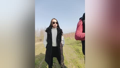 Video of a young woman with long brown hair and sunglasses, wearing a black trench coat over a white turtleneck, standing in a grassy field with a clear blue sky.