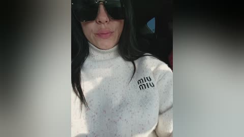 Video of a young woman with long black hair, wearing dark sunglasses, a white turtleneck sweater with \"Miu Miu\" printed in black, and a neutral background.