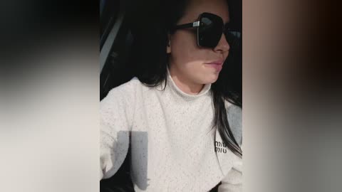 Video of a woman with long black hair, wearing large black sunglasses and a white turtleneck sweater, seated in a car, with a blurred, out-of-focus background.