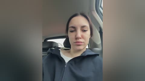 A video of a young woman with light skin, dark hair, and a neutral expression, wearing a black hoodie and headphones, sitting in a car.