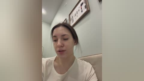 A video of a young woman with light skin and dark hair, wearing a white t-shirt, sitting indoors, with pastel walls and framed art behind her.