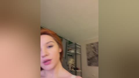 A blurry video of a woman with red hair, wearing pink lipstick, seen from the side in a dimly lit room with a white ceiling and grey walls.