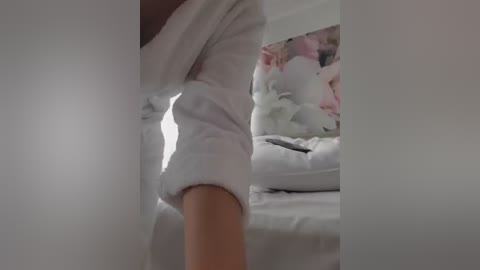 A video of a woman wearing a white, fluffy robe, standing in a bedroom with a white bed and pink and white floral-patterned pillows.
