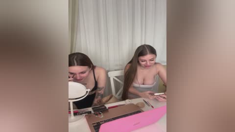 Video of two young women with long hair, wearing tank tops, sitting at a desk with makeup and tools, reflecting in a mirror.