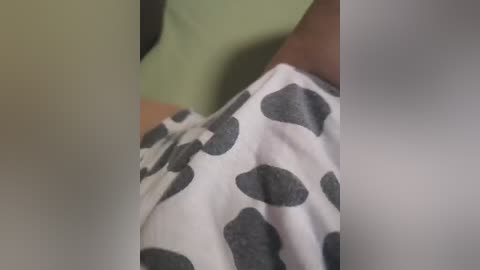 A close-up video of a person's arm in a white shirt with large gray cow patterns. The background shows a pale green wall. The focus is on the fabric texture and the shirt's unique design.