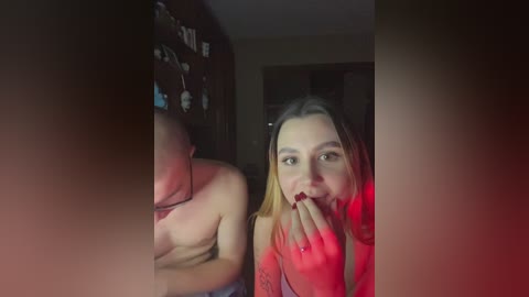 Video of a topless man with a muscular build, partially obscured by a red light, standing beside a young woman with blonde hair and red lipstick, in a dimly lit room with framed pictures on the wall.