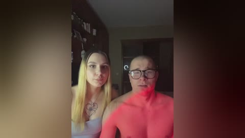 Video of a young woman with long blonde hair and a heart tattoo, and a middle-aged man with glasses, both topless, in a dimly lit room.