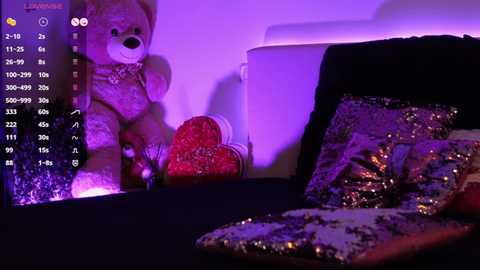 Video of a plush teddy bear, a red heart-shaped pillow, and a sequined cushion on a dark couch, bathed in purple light, set against a white wall.